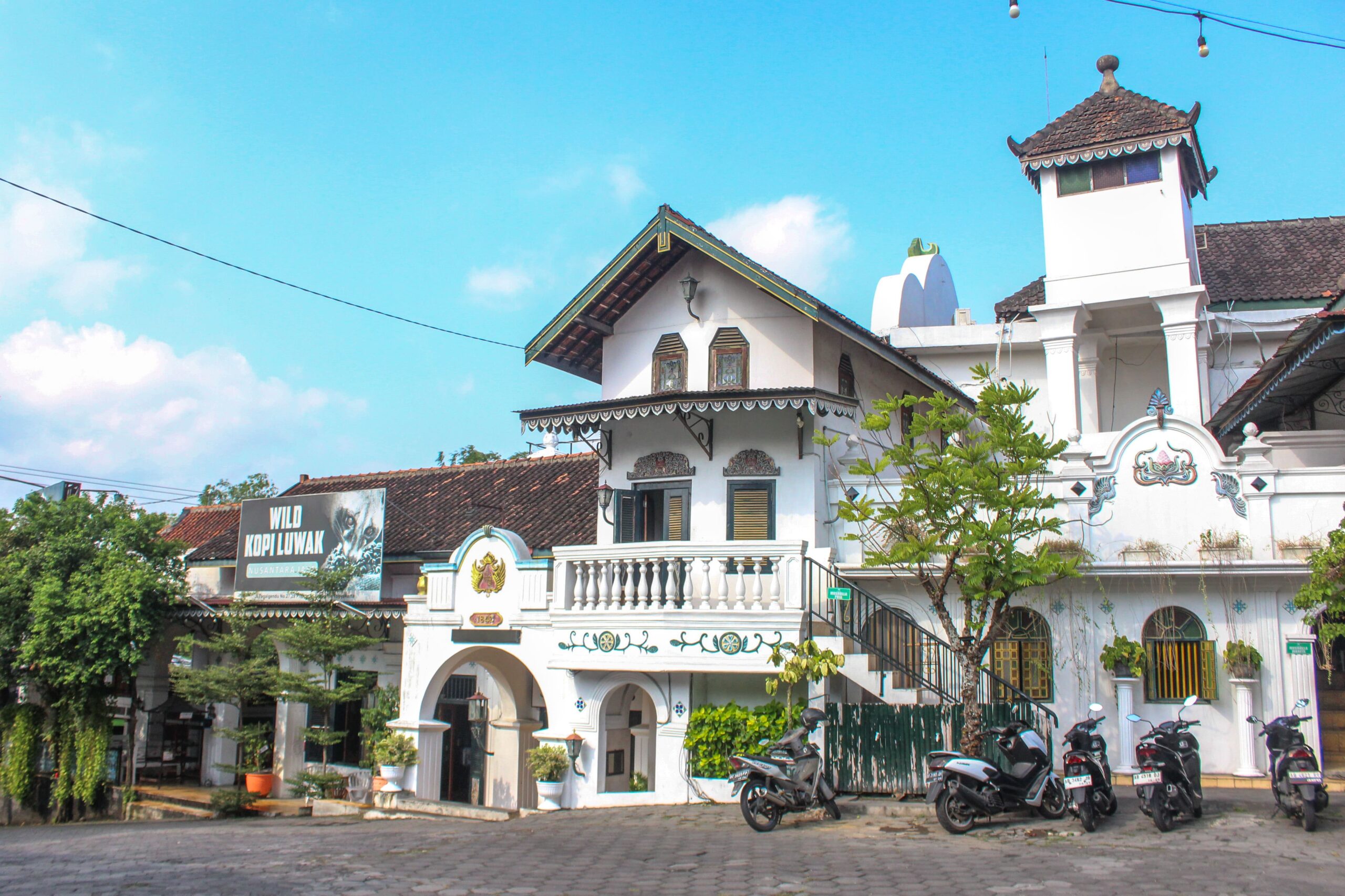 White colonial-style building with arches and balconies, motorcycles parked along the curb on a sunny street.