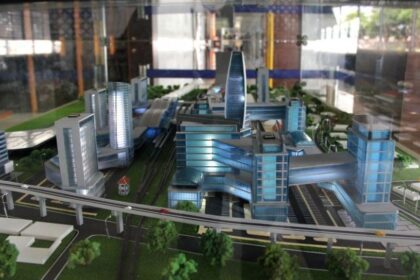 Scale model of a modern city with blue glass buildings, elevated train tracks, and green spaces nearby.