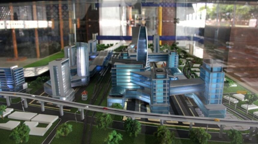 Scale model of a modern city with blue glass buildings, elevated train tracks, and green spaces nearby.