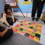 Two scouts sit on the floor playing a colorful tile game about Javanese script; a poster reads 'AYO SINAU AKSARA JAWA' nearby.
