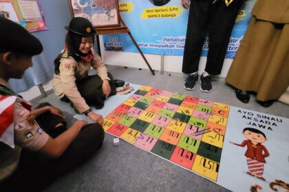 Two scouts sit on the floor playing a colorful tile game about Javanese script; a poster reads 'AYO SINAU AKSARA JAWA' nearby.