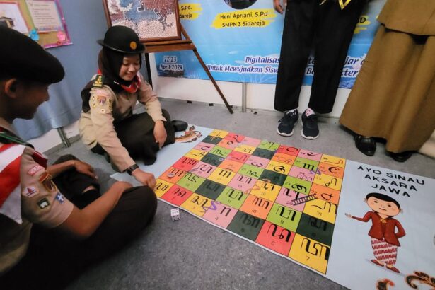 Two scouts sit on the floor playing a colorful tile game about Javanese script; a poster reads 'AYO SINAU AKSARA JAWA' nearby.