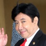 Man in a black suit and red tie waves hello indoors, smiling at the camera with a lapel pin visible on the jacket.