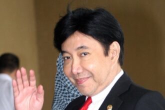 Man in a black suit and red tie waves hello indoors, smiling at the camera with a lapel pin visible on the jacket.
