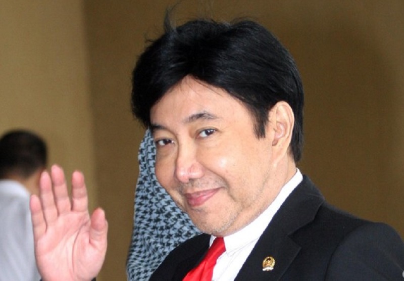 Man in a black suit and red tie waves hello indoors, smiling at the camera with a lapel pin visible on the jacket.