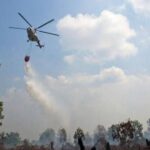Helicopter water bomber releasing a bucket to drop water on a smoky forest fire beneath a blue sky.