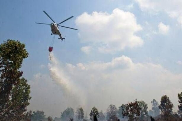 Helicopter water bomber releasing a bucket to drop water on a smoky forest fire beneath a blue sky.