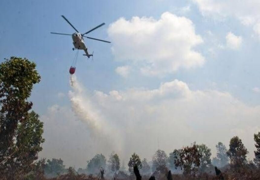 Helicopter water bomber releasing a bucket to drop water on a smoky forest fire beneath a blue sky.