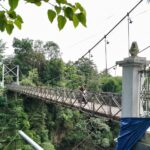 Person riding a motorcycle across a narrow suspension bridge over a deep, green forested gorge; ornate railing visible in the foreground.
