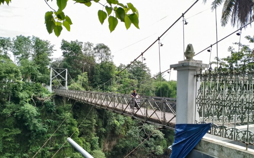 Person riding a motorcycle across a narrow suspension bridge over a deep, green forested gorge; ornate railing visible in the foreground.