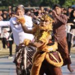 Dancer in a gold-brown lion costume with ornate headpiece performs at an outdoor cultural festival, surrounded by batik-clad spectators and fellow dancers.