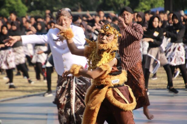 Dancer in a gold-brown lion costume with ornate headpiece performs at an outdoor cultural festival, surrounded by batik-clad spectators and fellow dancers.