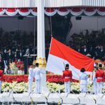 Indonesian flag ceremony with guards in white and red uniforms and a large seated audience in the background.