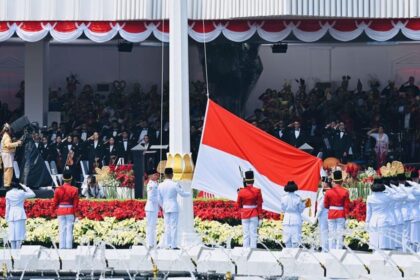 Indonesian flag ceremony with guards in white and red uniforms and a large seated audience in the background.