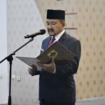 Man in a dark suit and traditional hat reading from a folder at a microphone podium in a formal setting.