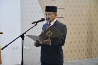 Man in a dark suit and traditional hat reading from a folder at a microphone podium in a formal setting.