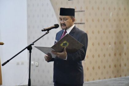 Man in a dark suit and traditional hat reading from a folder at a microphone podium in a formal setting.