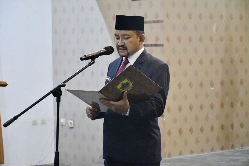 Man in a dark suit and traditional hat reading from a folder at a microphone podium in a formal setting.