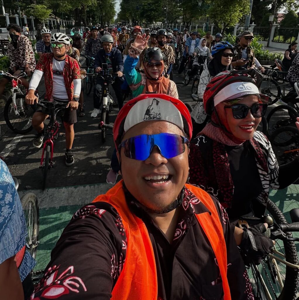 698b06ee-f91d-4167-af47-501b63776d7a - Mabur.co Smiling rider in an orange safety vest takes a selfie with a large group of cyclists behind him at an outdoor ride.