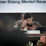 Indonesian finance ministry official in a batik shirt, head in hand at a formal meeting; nameplate reads Purbaya Yudhi Sadewa.