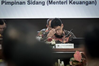 Indonesian finance ministry official in a batik shirt, head in hand at a formal meeting; nameplate reads Purbaya Yudhi Sadewa.