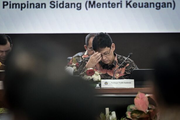 Indonesian finance ministry official in a batik shirt, head in hand at a formal meeting; nameplate reads Purbaya Yudhi Sadewa.