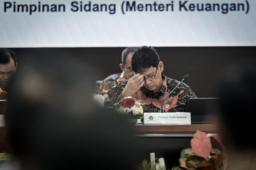 Indonesian finance ministry official in a batik shirt, head in hand at a formal meeting; nameplate reads Purbaya Yudhi Sadewa.