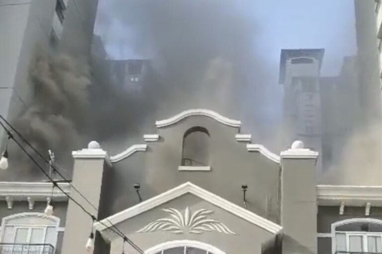 Gray ornate building in the foreground with thick black smoke rising from behind it, indicating a fire in the background.