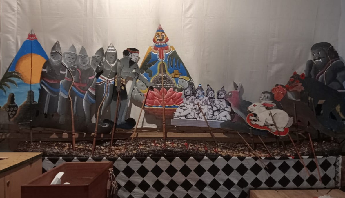 Colorful cutout puppets arranged on a stage, depicting a mythological scene with deities and warriors, with rods in front for manipulation.