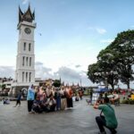 Group of people posing for a photo in a public square near a tall white clock tower with a pointed roof, while a photographer kneels to capture the shot.