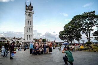 Group of people posing for a photo in a public square near a tall white clock tower with a pointed roof, while a photographer kneels to capture the shot.