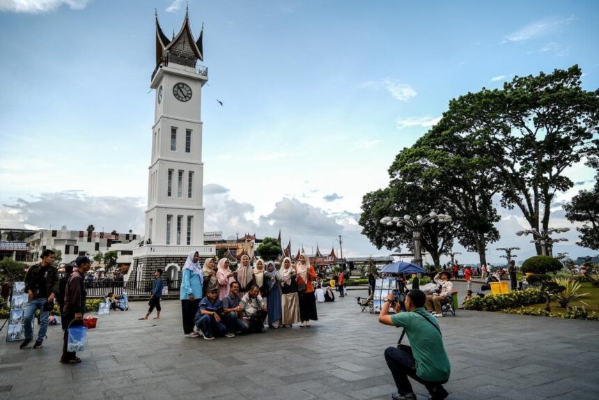 6a1e95e8a023b564450955 - Mabur.co Group of people posing for a photo in a public square near a tall white clock tower with a pointed roof, while a photographer kneels to capture the shot.