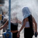 Two women in summer clothes stand near a street sprinkler, misting around them on a hot day.