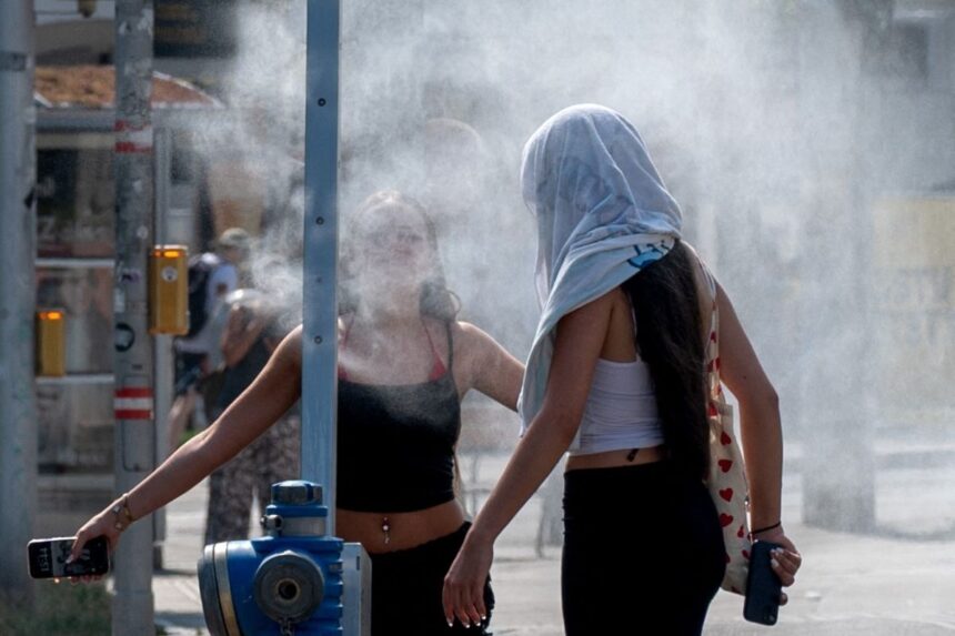6a44ed5e0fb76 - Mabur.co Two women in summer clothes stand near a street sprinkler, misting around them on a hot day.