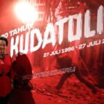 Smiling woman in a red outfit posing beside a large red banner with white text and dates.