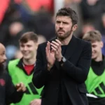 Man in a dark coat claps on a football pitch, with players in green bibs behind him.