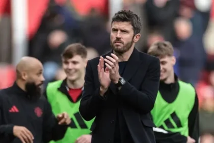 Man in a dark coat claps on a football pitch, with players in green bibs behind him.