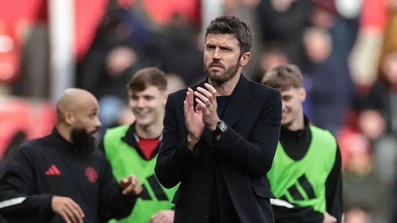 Man in a dark coat claps on a football pitch, with players in green bibs behind him.