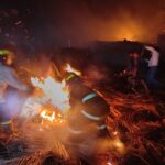 Nighttime scene of firefighters in turnout gear fighting a blaze, sparks flying around as they douse the flames with a hose.