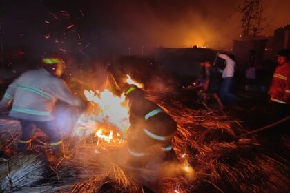 Nighttime scene of firefighters in turnout gear fighting a blaze, sparks flying around as they douse the flames with a hose.