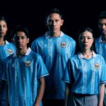 Five young adults pose together in matching light-blue soccer jerseys with a crest, against a dark background.