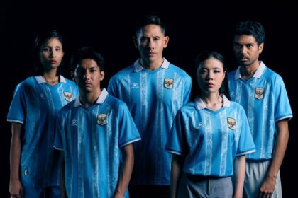Five young adults pose together in matching light-blue soccer jerseys with a crest, against a dark background.