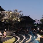 Audience seated around a circular stage for an outdoor performance at dusk, with temple buildings in the background.