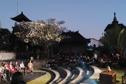 Audience seated around a circular stage for an outdoor performance at dusk, with temple buildings in the background.