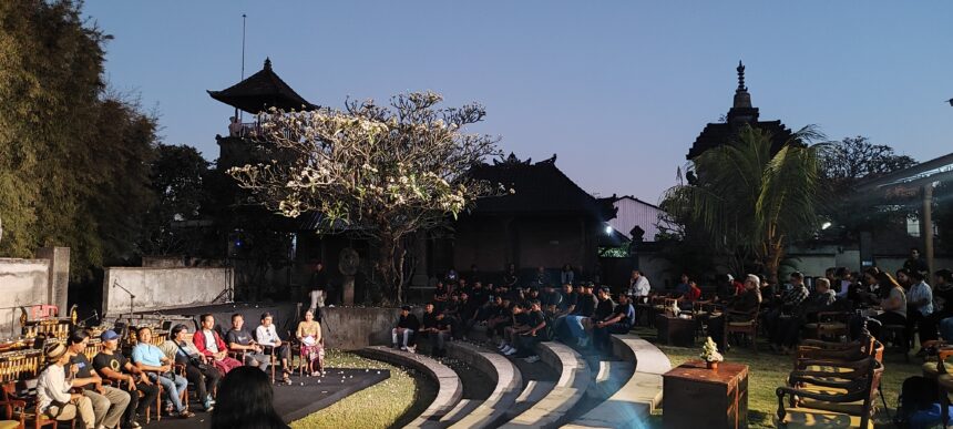Audience seated around a circular stage for an outdoor performance at dusk, with temple buildings in the background.