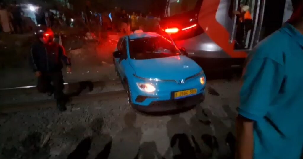 Blue compact car with front-end damage on a dark street, red emergency lights flashing in the background, bystanders nearby.