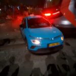 Blue compact car with front-end damage on a dark street, red emergency lights flashing in the background, bystanders nearby.