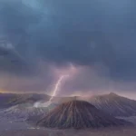 Volcanic crater landscape under stormy clouds with a bright lightning bolt striking the crater area at dusk.