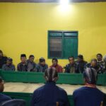 Group of men in traditional attire sit along a green rectangular barrier in a bright yellow room, with a camera on a tripod to the left.