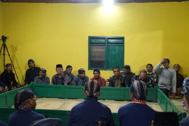 Group of men in traditional attire sit along a green rectangular barrier in a bright yellow room, with a camera on a tripod to the left.